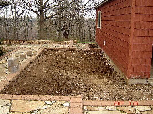An empty, rectangular garden bed with brick border next to a red-sided house.
