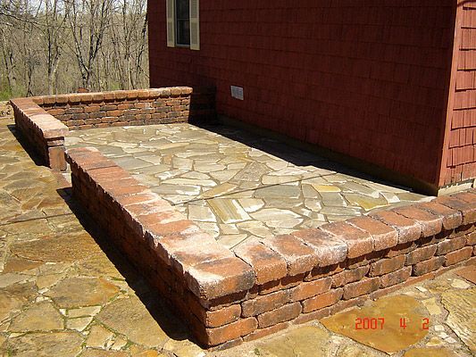 Brick-bordered flagstone patio next to a red house, surrounded by trees.