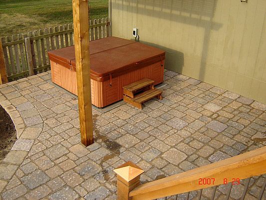 Hot tub on a patterned stone patio, wooden steps, and a brown cover.