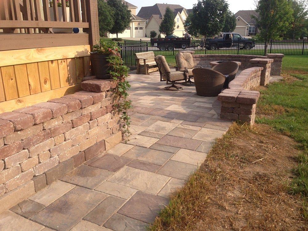 Patio with brick walls and paving stones; seating area with chairs, bordering a deck and grass.