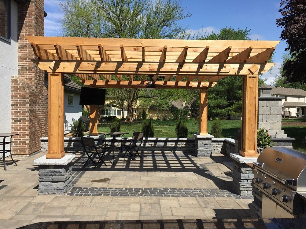 Wooden pergola over a patio with outdoor seating, a grill, and a brick house in the background.