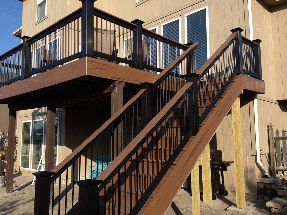 Deck with brown planks and black railings, leading to a second-story level; exterior of a house.