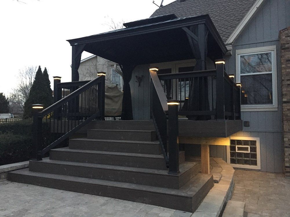 Dark deck with stairs and pergola, lit by small lights, attached to a gray house.