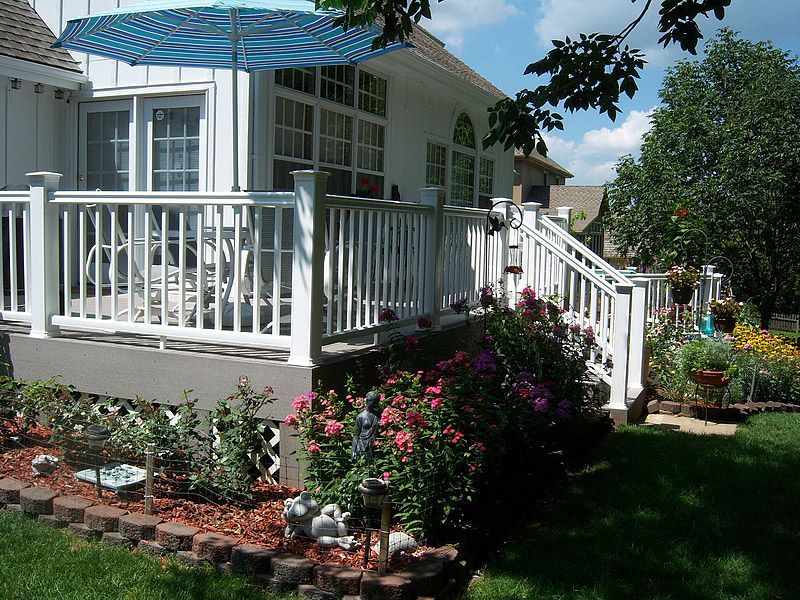 White deck with white railing, flowers, umbrella, and a house.