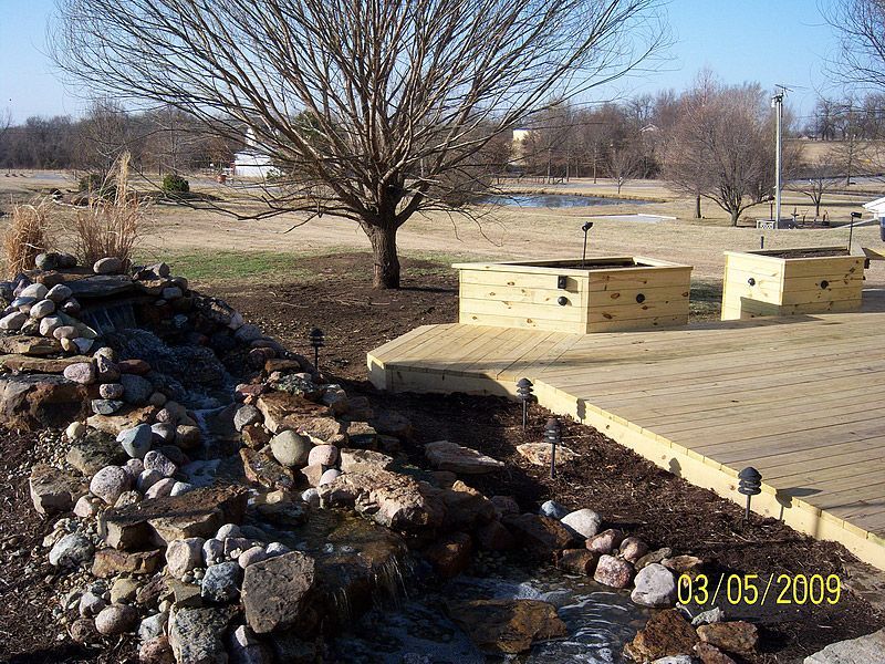 Landscaped yard with waterfall, deck, and two wooden structures. Bare tree, brown grass, and blue sky.
