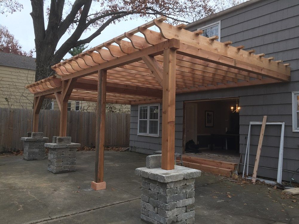 Wooden pergola attached to a house, supported by stone pillars.
