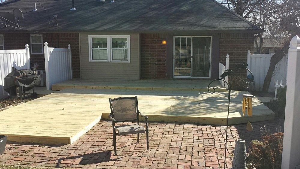 Backyard with brick patio, unfinished wooden deck, white fence, and house with window and sliding door.