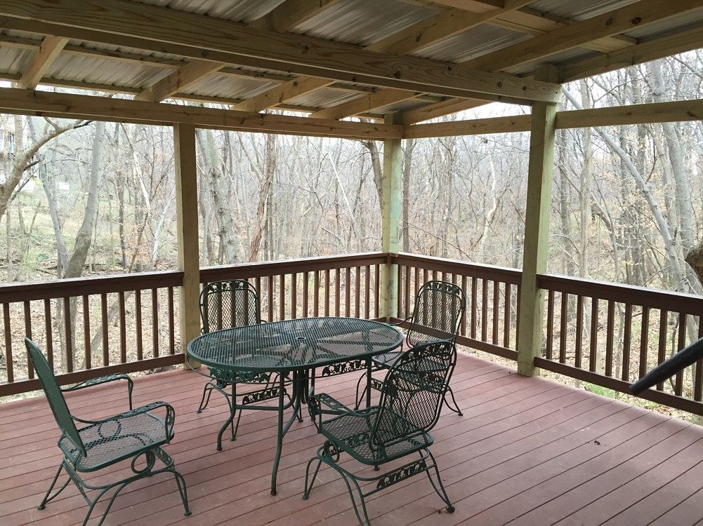Covered deck with a metal table and chairs, overlooking a wooded area.