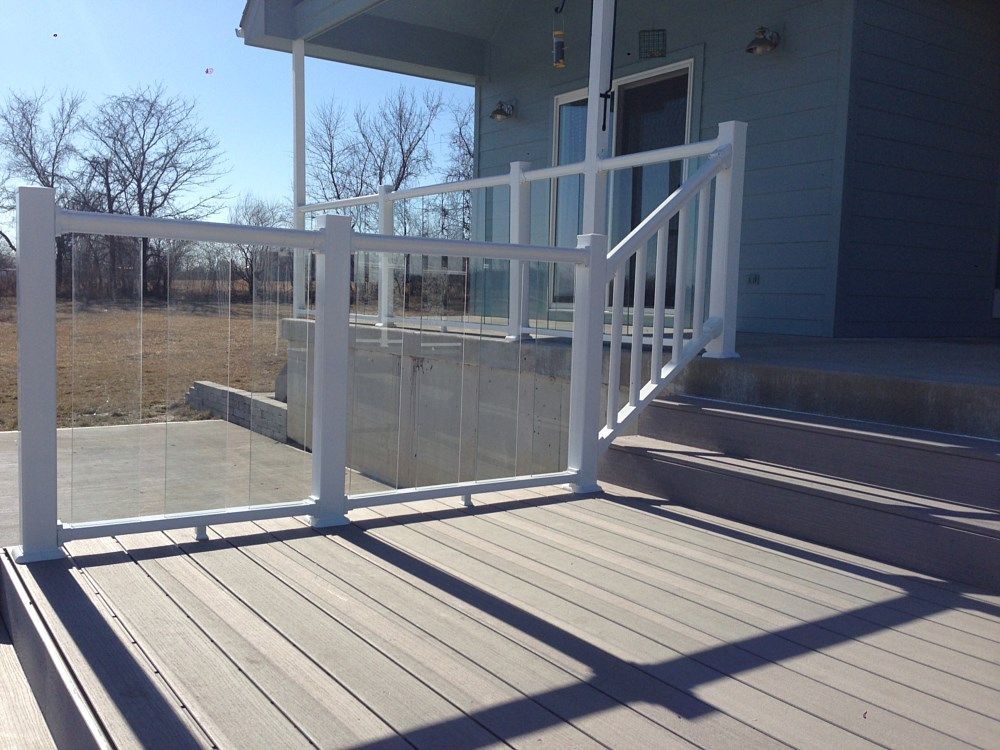 White railing with glass panels on a wooden deck, leading to steps and a light blue house.