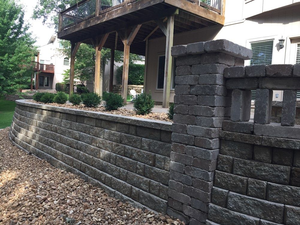 Retaining wall built of gray blocks, topped with shrubs, beneath a wooden deck and house.