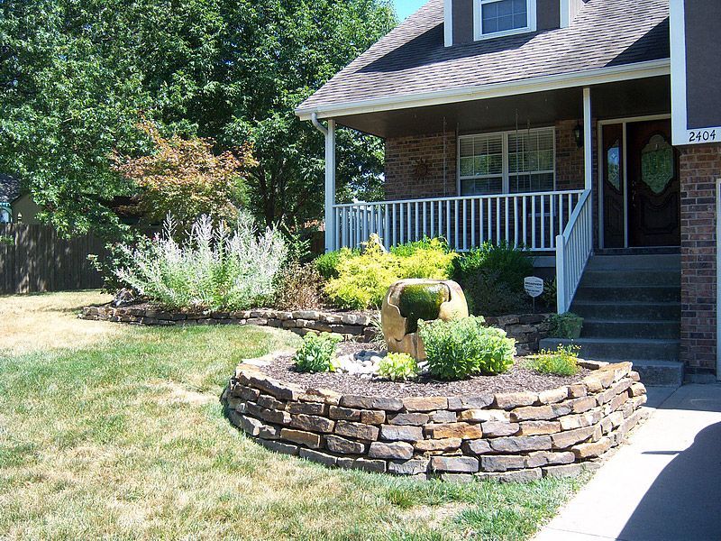 House with stacked stone garden bed, water fountain, and porch. Green lawn and trees surround.
