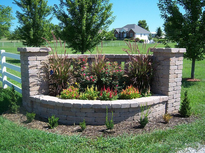 Stone planter with flowers, flanked by stone columns and white fence; a house is in the background.