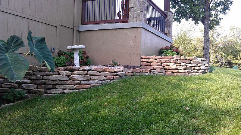 Stone retaining walls border a yard with green grass, leading to a porch and house exterior.