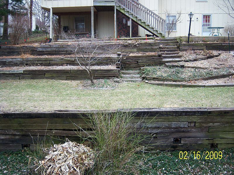 Wooden retaining walls terraced into a hillside, leading to a house with a deck and stairs.