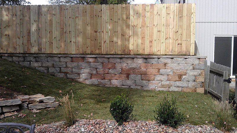 Wooden fence atop a stone retaining wall in a yard with grass and bushes.