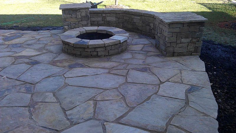 Stone patio with fire pit and curved stone seating area, surrounded by grass.