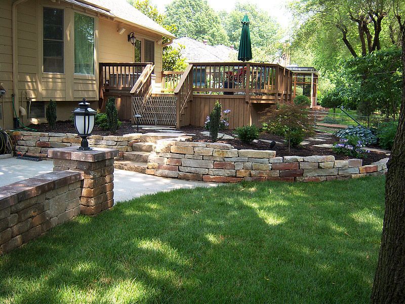 Backyard with tiered stone walls, wooden deck, green lawn, and trees.