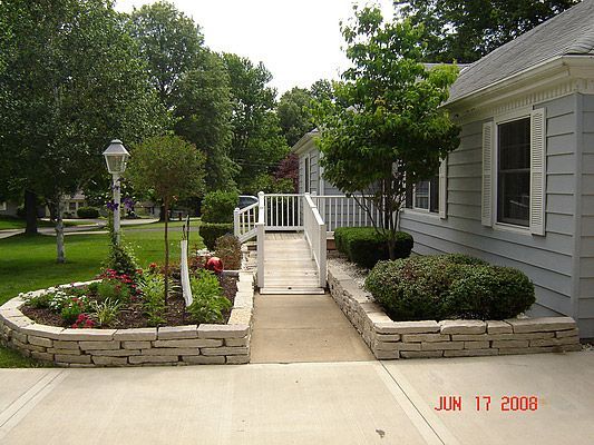 A residential house entrance with a ramp, flanked by stone-lined flowerbeds with greenery.