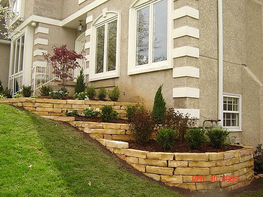Stone retaining walls with plants and grass leading to a two-story house with white trim.