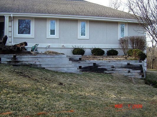 Gray house with tiered, weathered wooden retaining wall in front, on a grassy hill.