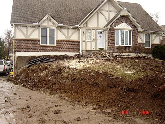 House with excavated front yard, showing exposed foundation and driveway covered in mud.