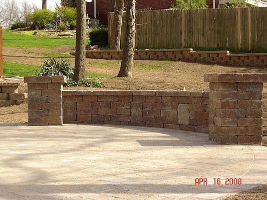 Stone retaining wall with pillars, bordering a concrete patio. Trees and a wooden fence are visible in the background.