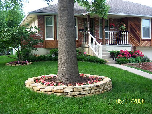 Tree trunk surrounded by stone-walled flower bed in front of a house with a brick and wood exterior, green grass, and blooming flowers.