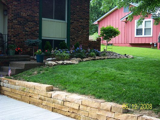 A brick building with a stone retaining wall and a grassy yard. A pink house sits in the background.