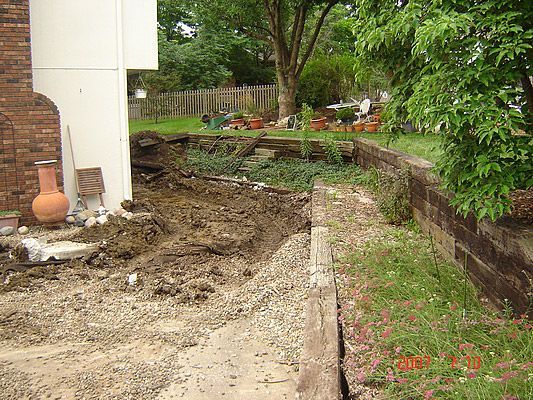 Yard with a collapsed retaining wall, exposing dirt and debris next to a house and grassy area.
