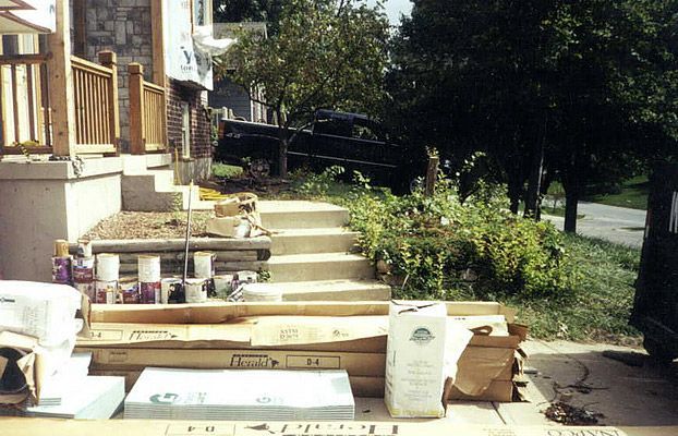 Materials and debris piled on front steps of a house; wooden porch railing, concrete steps, and a black fence.