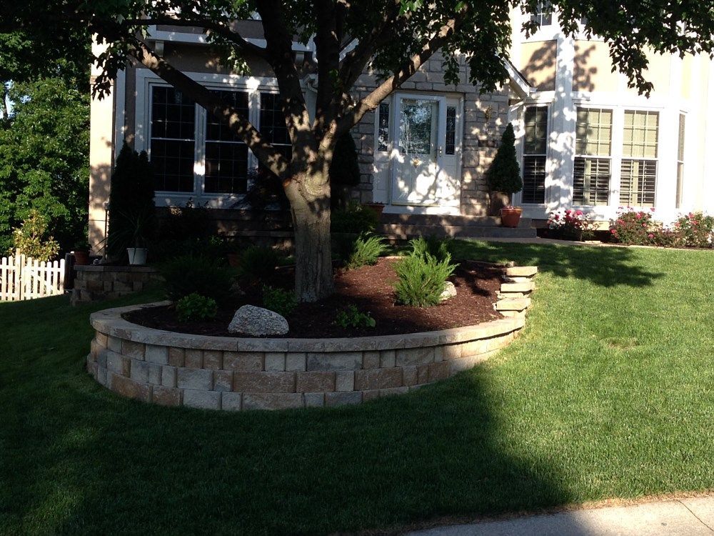 A house with a circular brick planter around a tree in front. Green grass and a sunny day.