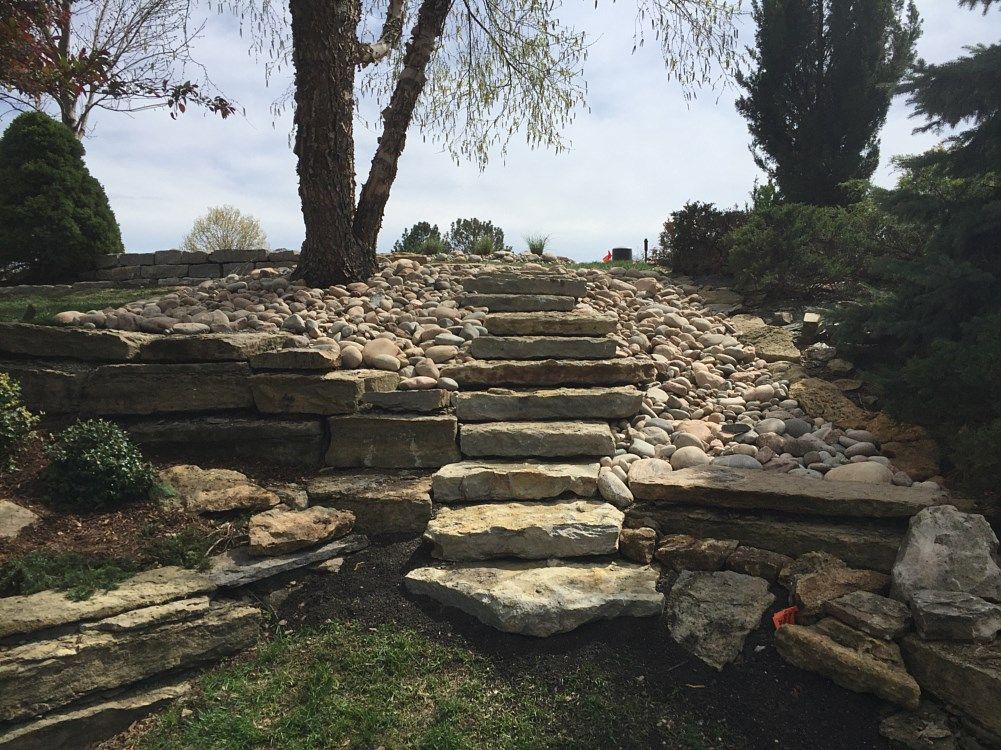 Stone steps ascend a rocky hillside, flanked by landscaping and a tree.