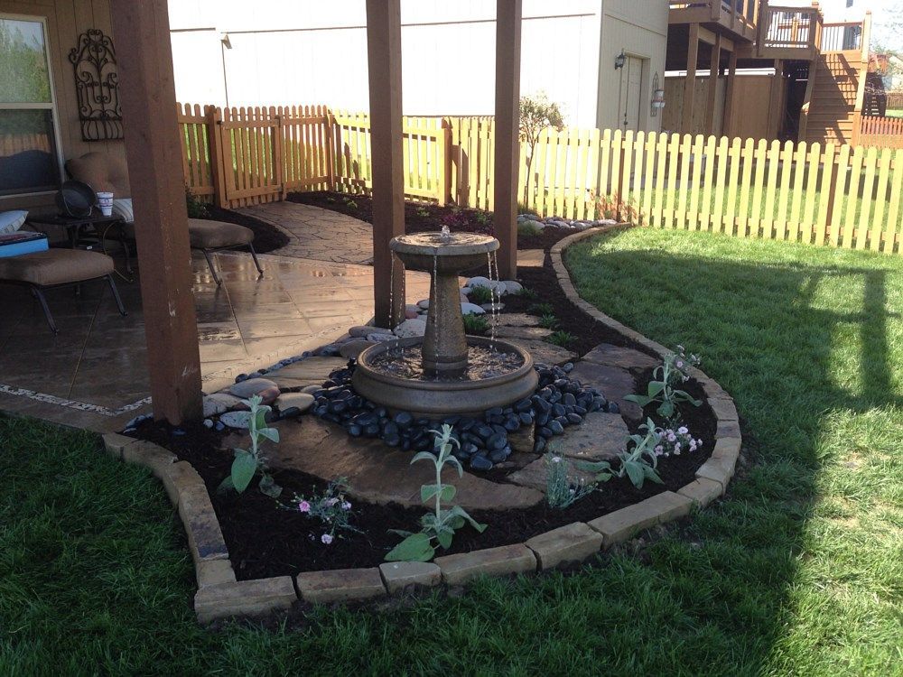 Backyard with a fountain, stone walkway, and small flower garden, beside a patio.
