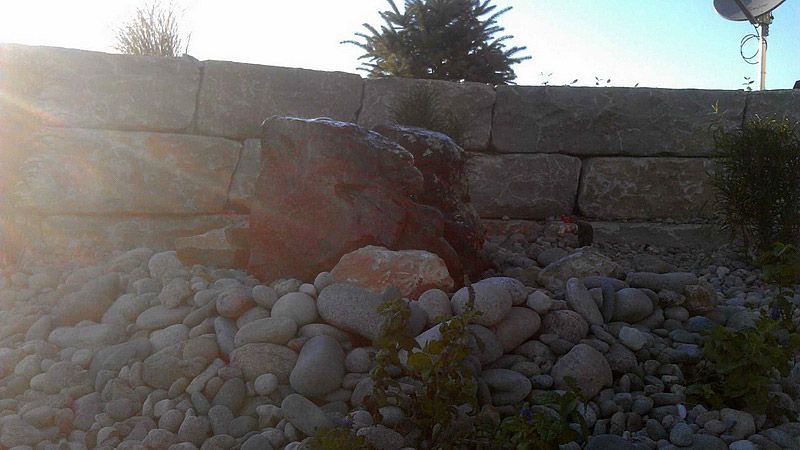 A rock water fountain with a stone retaining wall behind it, set in a sunny outdoor setting.