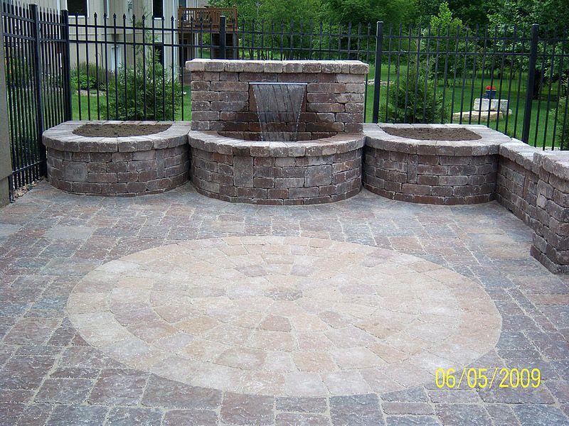 Stone water feature with three basins on a brick patio with an inlaid circle, next to a black fence.