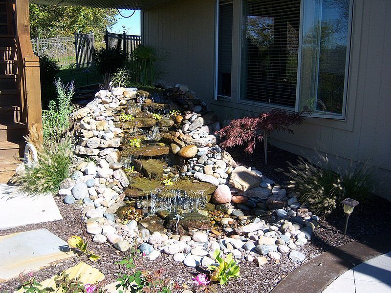Rock waterfall feature with plants and pebbles next to a house.