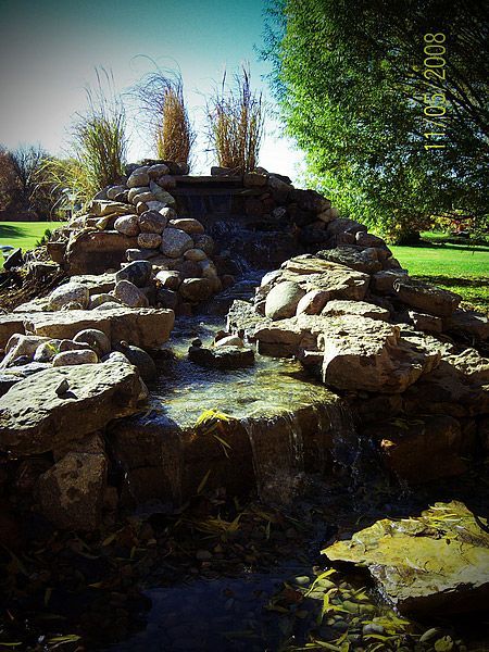 Rock waterfall feature with flowing water and tall grasses. Green lawn and tree in background.