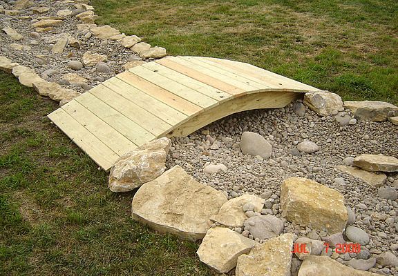 Wooden arched bridge over a dry creek bed with large rocks and gravel.