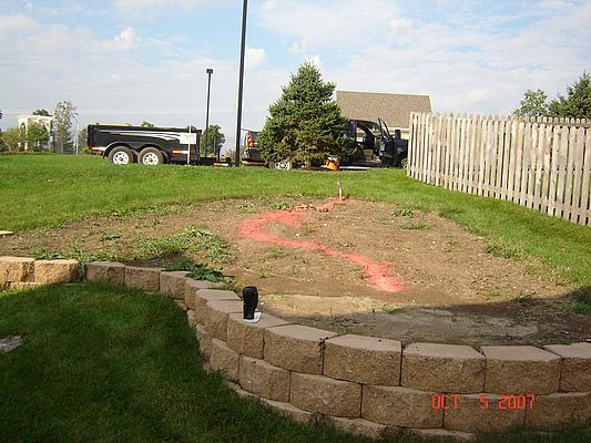 A sloped backyard with a retaining wall and marked garden bed near a fence and trailer.