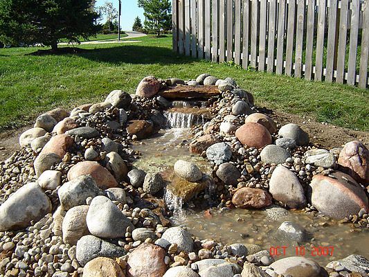 Small rock waterfall feature in a yard with grass and a wooden fence.
