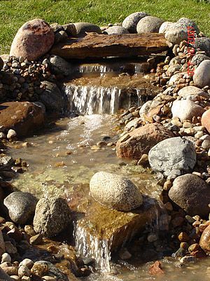 Small waterfall feature cascading over rocks into a pool, surrounded by stones and grass.
