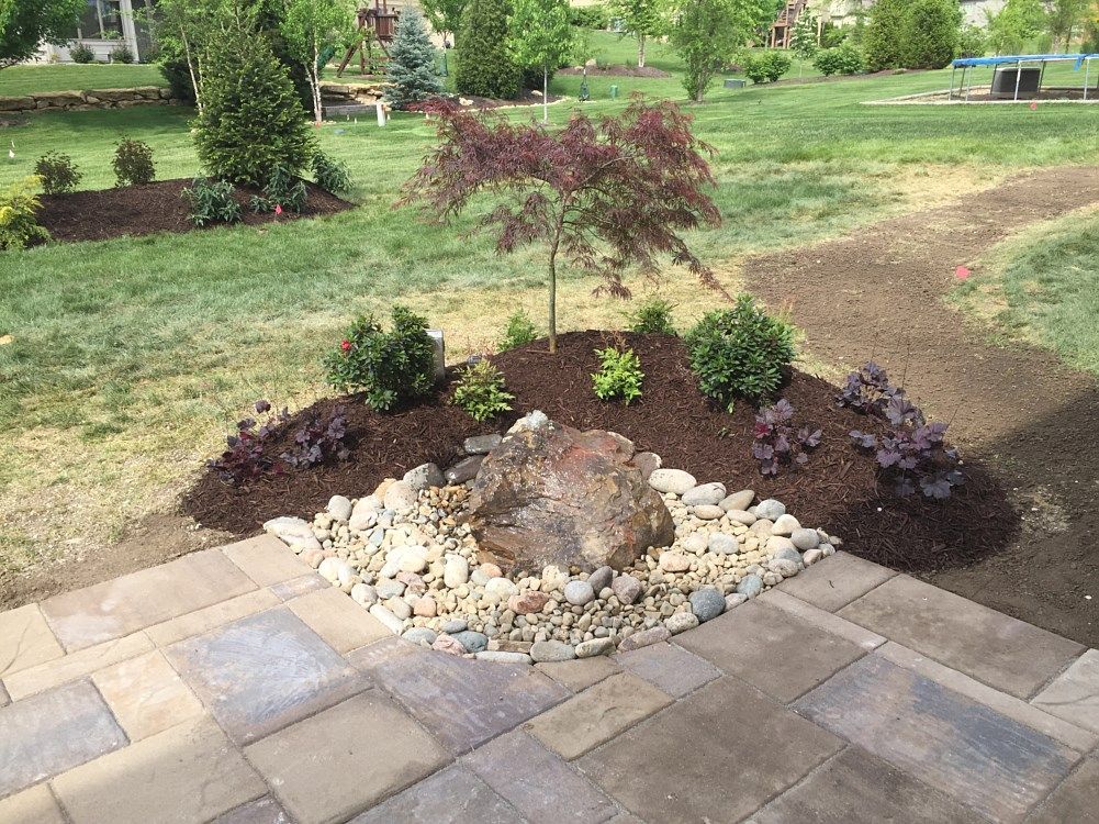 Rock fountain surrounded by plants and stones on a stone patio with a grassy backyard.
