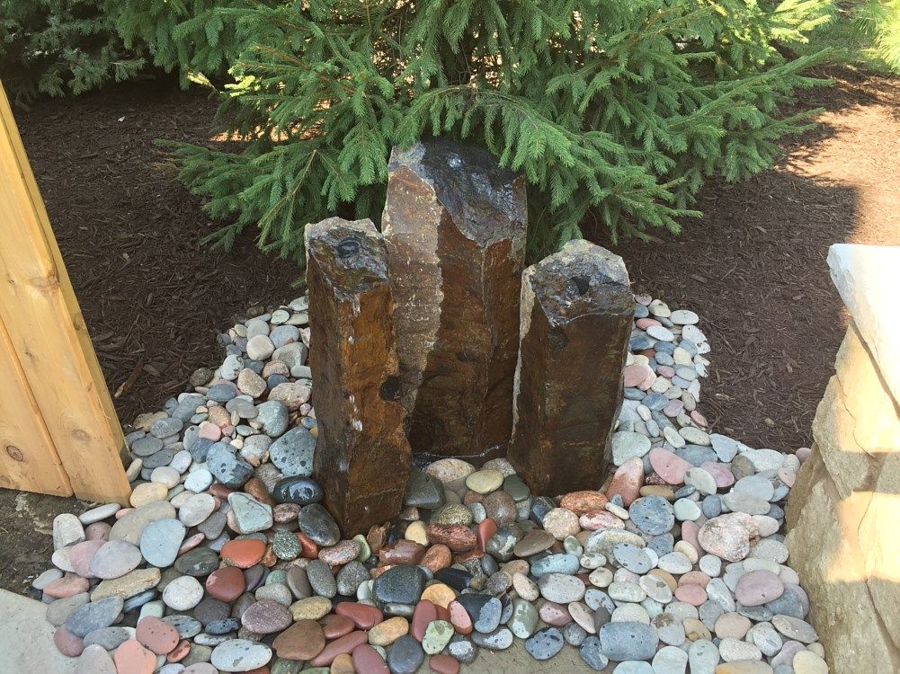 Water fountain with three dark stone columns surrounded by colorful river rocks.