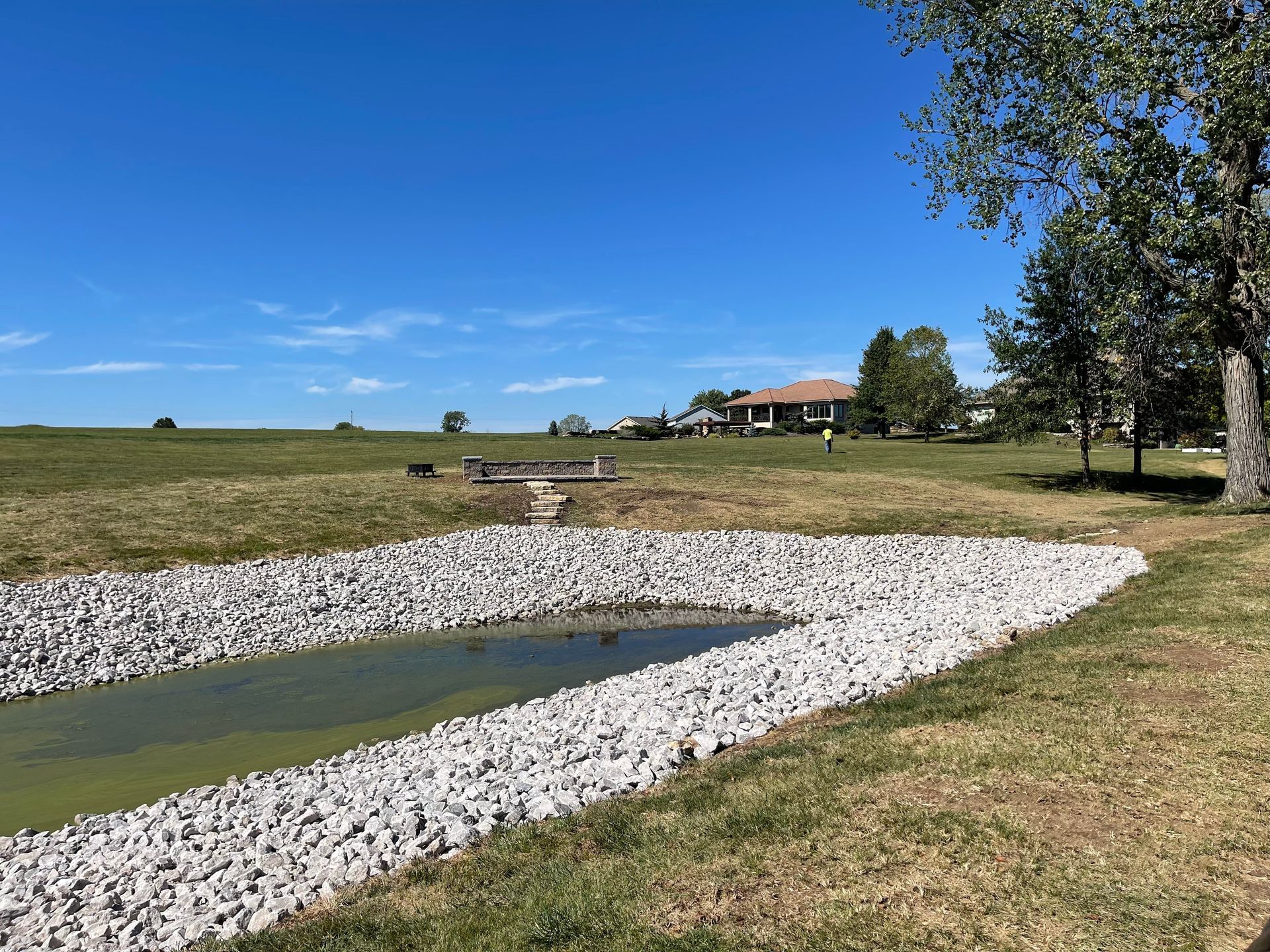 White rock-lined pond in a grassy field with a clear blue sky and distant house.