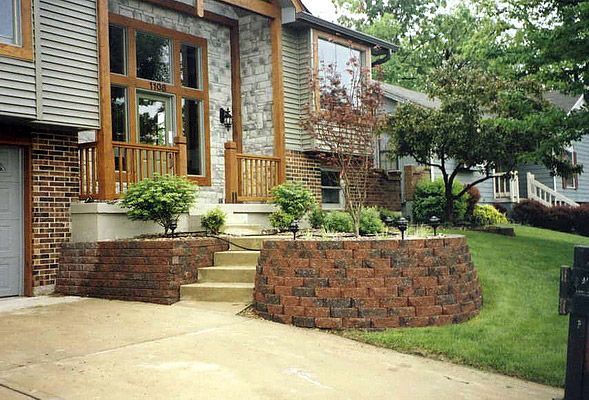 Brown brick retaining wall and steps leading to a house with a wooden door frame, surrounded by grass and landscaping.