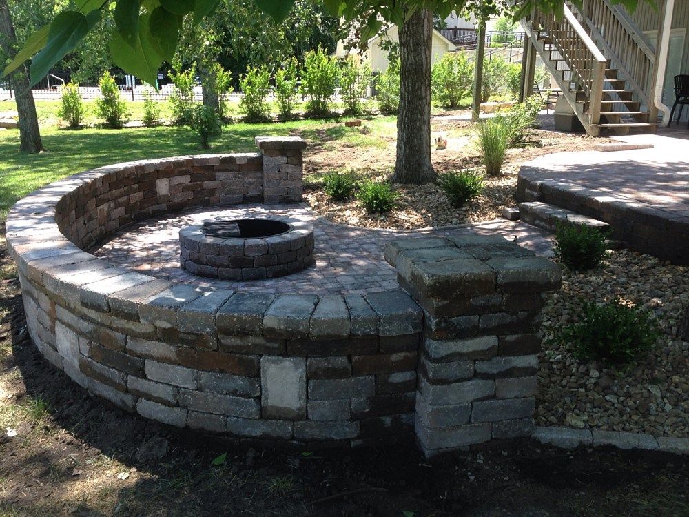 A stone fire pit surrounded by a curved wall, near a tree and deck.