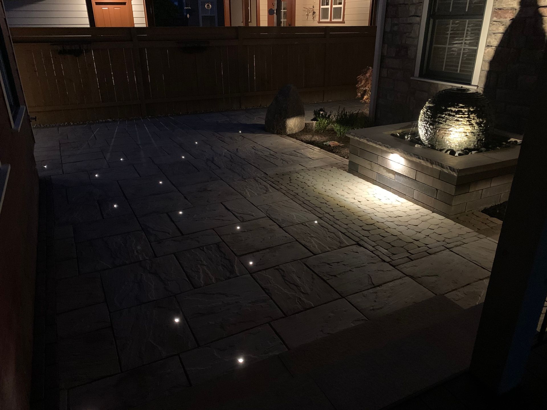 Dark stone patio at night with small ground lights and a lit fountain beside a building entrance