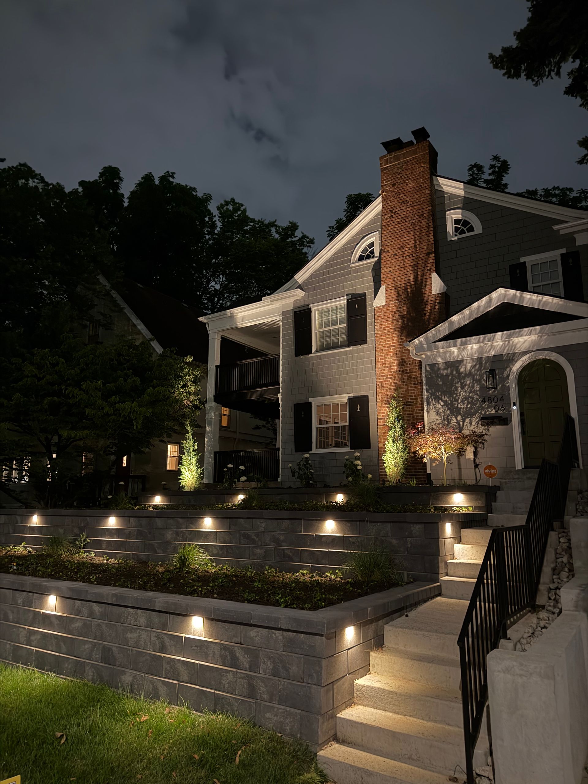 Nighttime view of a lit stone house with garden steps, brick chimney, and trees against a dark sky