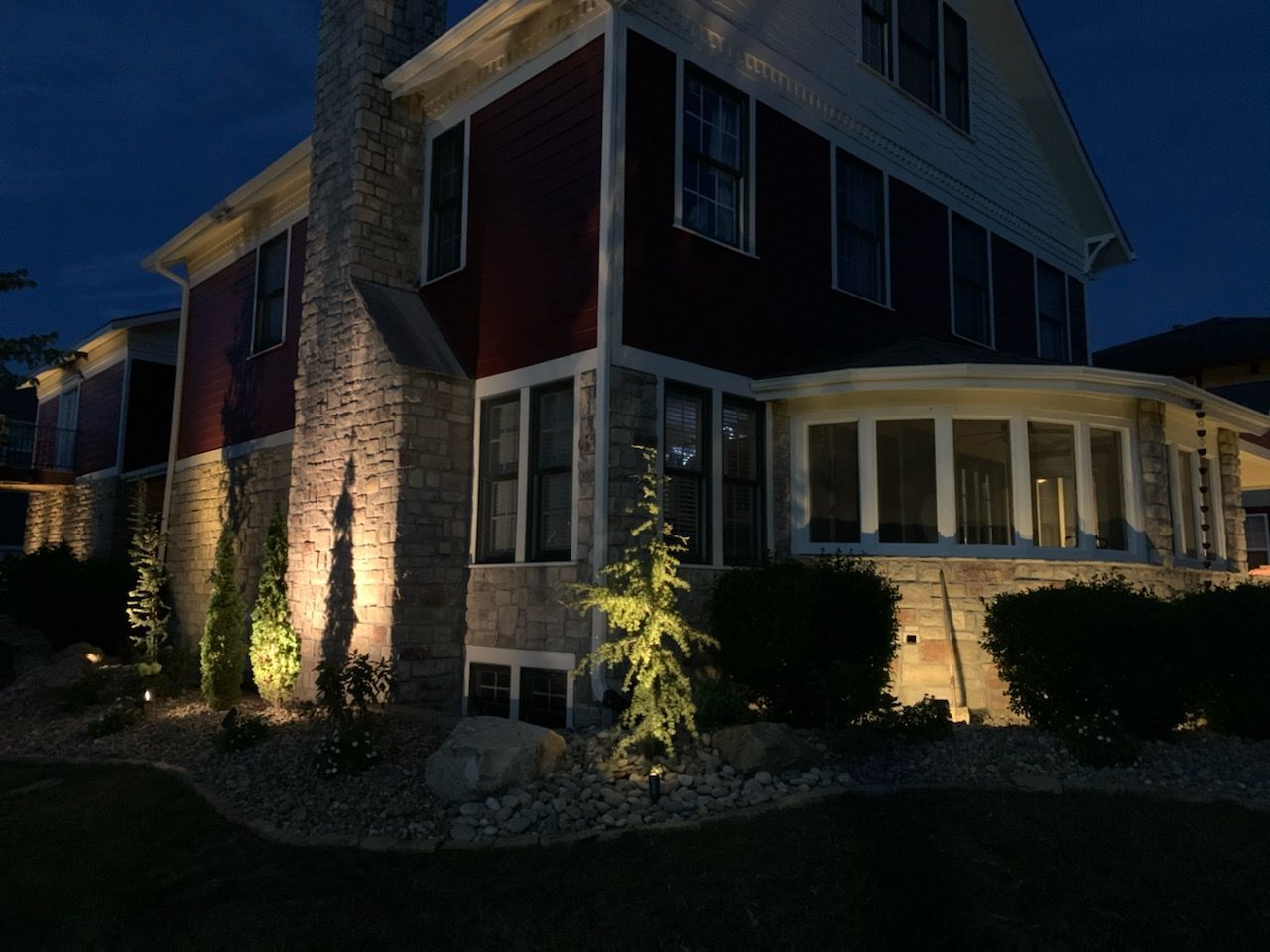 Dusk-lit two-story house with stone facade, red siding, and landscaped bushes beside a sidewalk