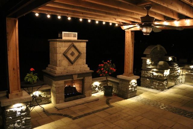 Covered outdoor patio with lit brick fireplace, wooden beams, and decorative stone planters at night